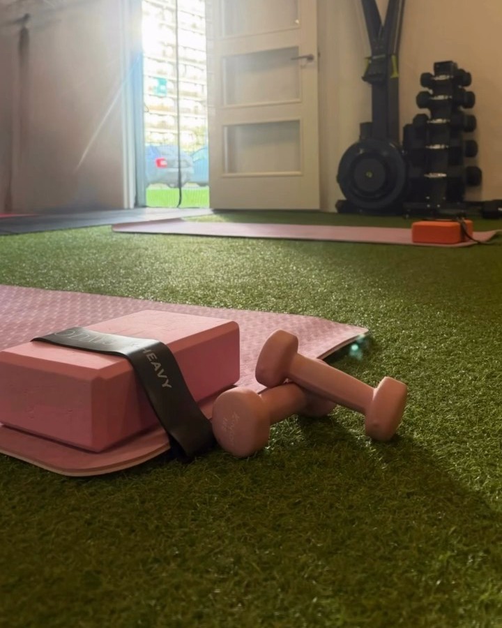 Een roze blok, weerstandsband en dumbbells op een mat bij de open deur van de studio